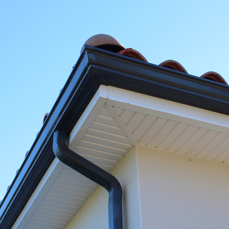 Gouttière noire et soffite blanc pour un coin de toit Coin de toit avec gouttière noire et descente, installé sur un soffite ventilé en vinyle blanc et mur beige. Tuiles de terre cuite.