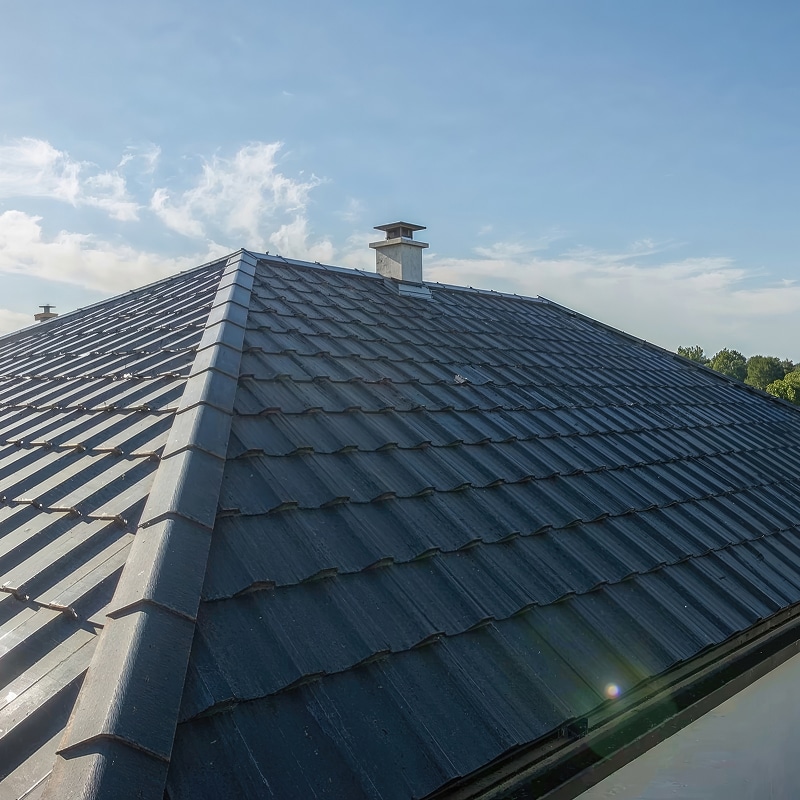 Tuiles métalliques : pose et rénovation de toiture sombre. Toiture sombre et étendue en tôle profilée (aspect tuile) avec faîtage. Une cheminée blanche se dresse contre le ciel bleu et nuageux.